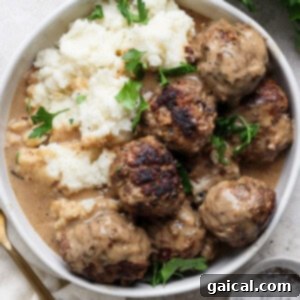 A plate of Minnesota Swedish Meatballs covered in gravy, served with mashed potatoes and garnished with fresh parsley. A fork and a small bowl of pepper sit adjacent to the plate, inviting you to dig in.