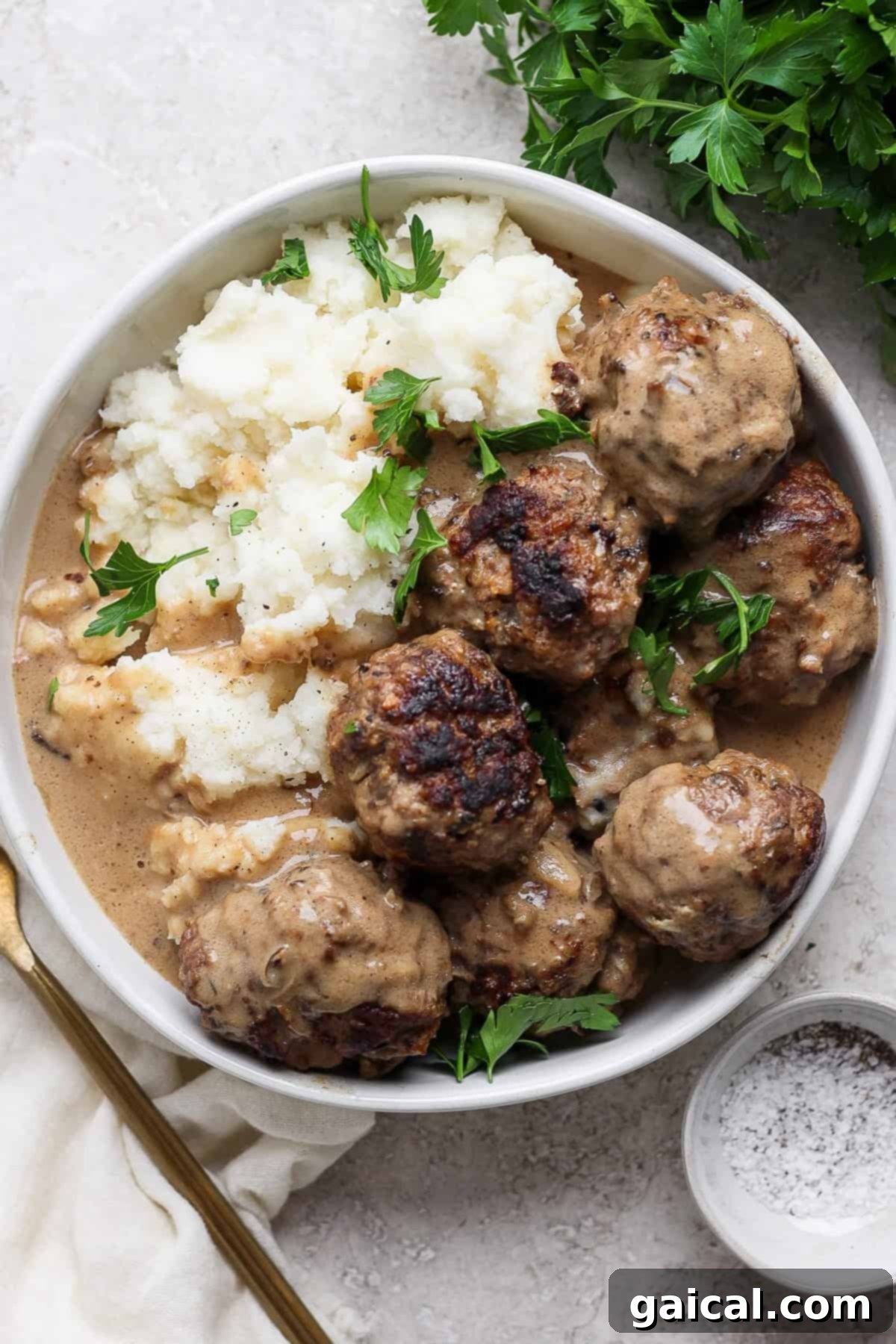 North Star State Swedish Meatballs 6 A beautifully plated dish of Minnesota Swedish Meatballs smothered in gravy, served alongside creamy mashed potatoes and garnished with vibrant fresh parsley. A fork and a small dish of pepper complete the inviting presentation.