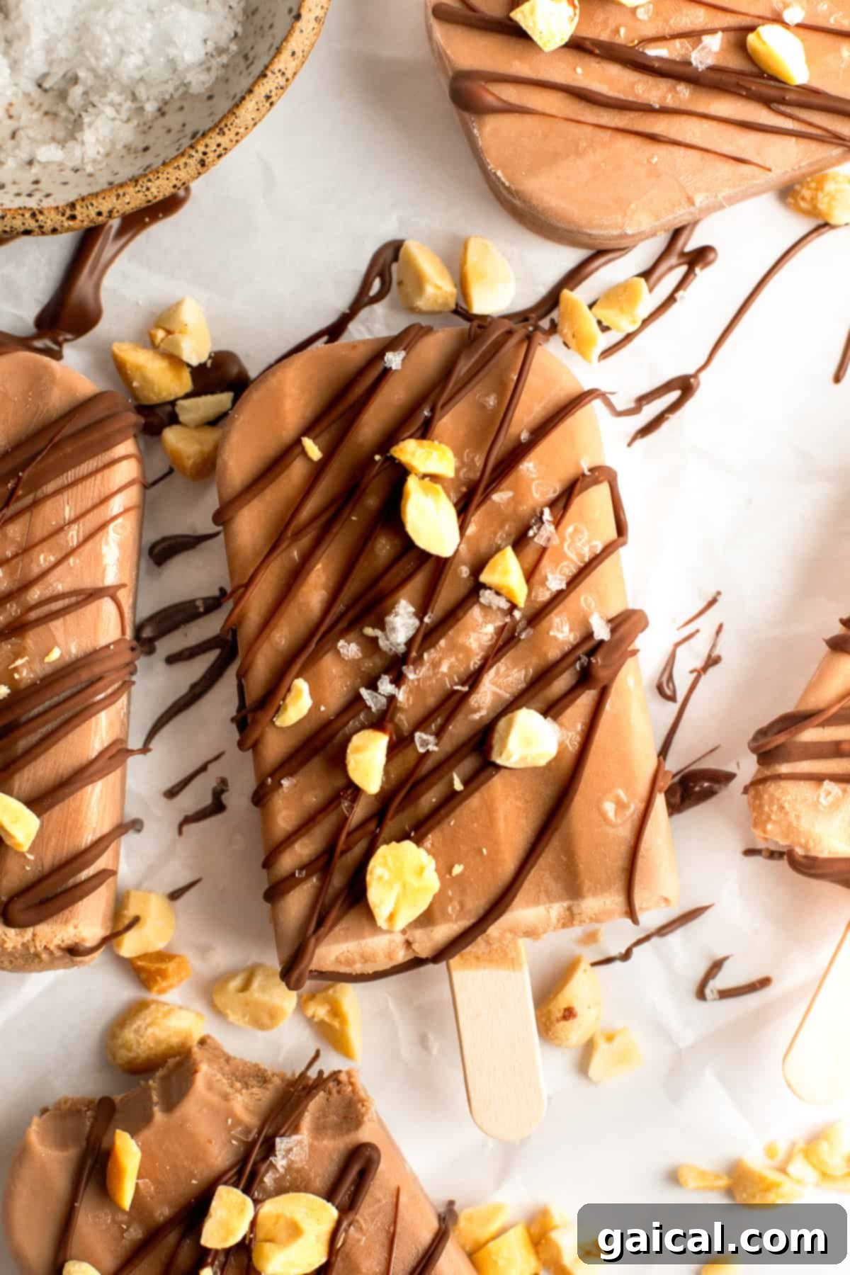 Guilt-Free Protein Fudge Pops 2 Chocolate popsicles drizzled with chocolate and sprinkled with chopped nuts and sea salt on a white surface.