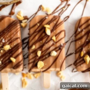 Three chocolate popsicles drizzled with dark chocolate and sprinkled with crushed nuts and sea salt are arranged on a parchment-covered surface.