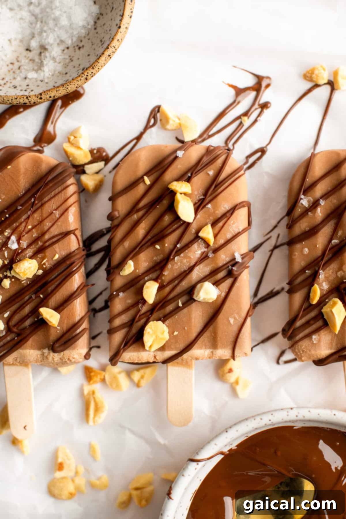 Guilt-Free Protein Fudge Pops 7 Three chocolate popsicles drizzled with dark chocolate and sprinkled with crushed nuts and sea salt are arranged on a parchment-covered surface.