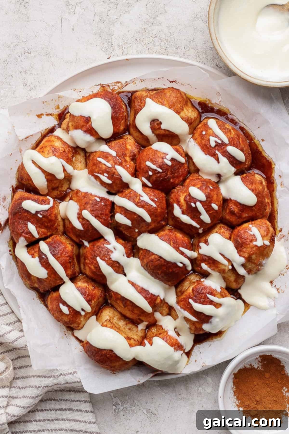 A golden-brown cinnamon pull-apart bread, generously topped with a creamy white glaze, rests on parchment paper. A small bowl of extra glaze and a sprinkle of cinnamon are nearby.
