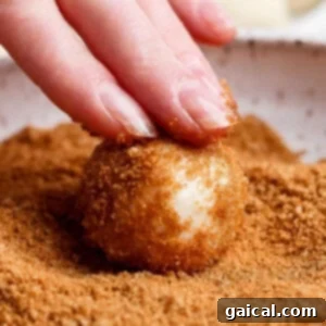A hand is actively rolling a dough ball in a bowl filled with a delicious brown sugar and cinnamon topping, ensuring an even coating.