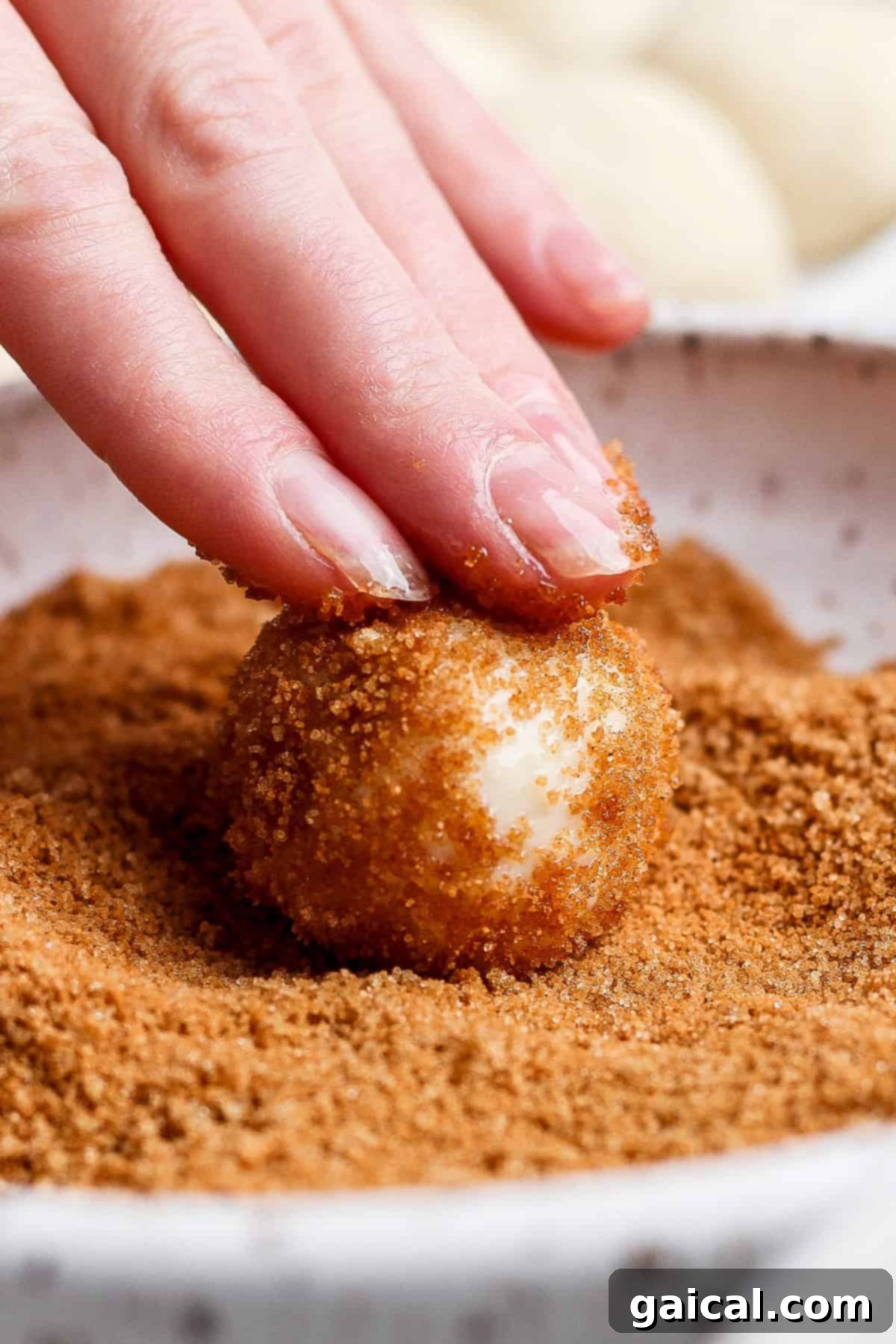 A close-up shot of a hand gently rolling a small dough ball through a bowl filled with brown sugar and cinnamon topping.