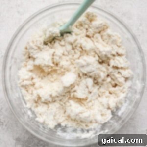 A clear bowl with a light blue spatula contains clumps of mixed flour and butter, forming a crumbly dough, on a textured gray surface.