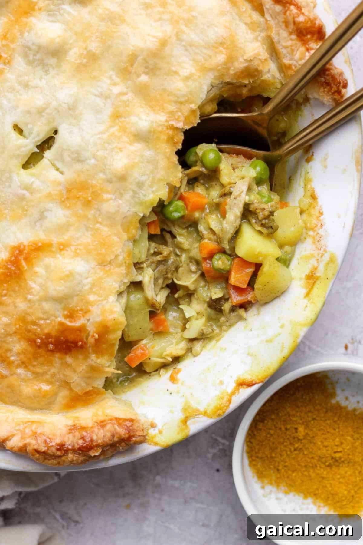 A pot pie with a flaky crust partially open, revealing a filling of chicken, carrots, peas, and potatoes. Two spoons are placed inside, next to a small dish of curry powder.