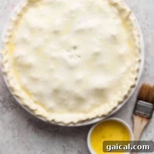 Unbaked pie with a crimped edge on a round dish, accompanied by a small bowl of egg wash and a pastry brush on a textured surface.