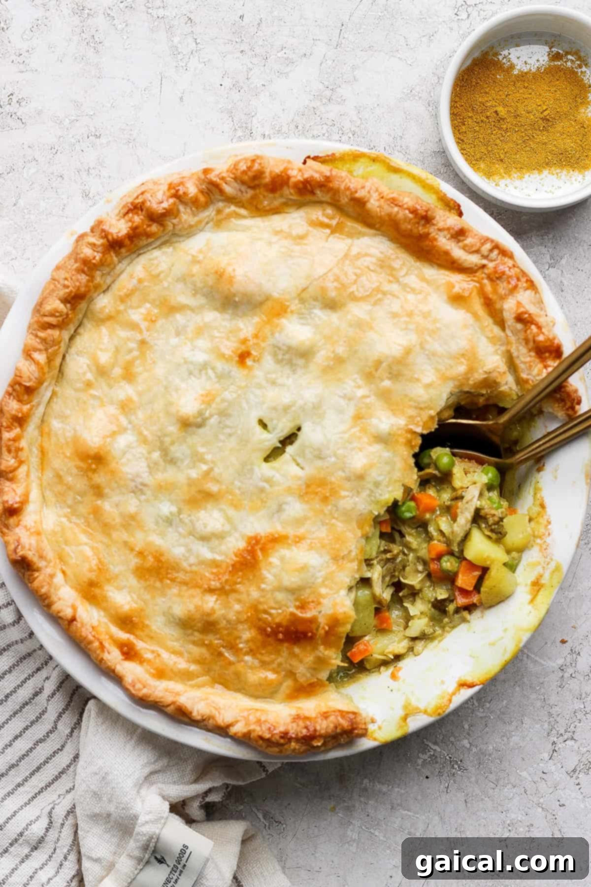 A savory pie with a golden crust is partially sliced, revealing a filling of vegetables and chicken. Two spoons are in the pie, and a bowl of seasoning is nearby.