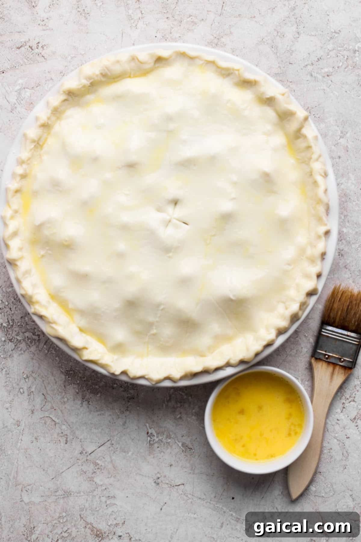 Unbaked pie with a crimped edge on a round dish, accompanied by a small bowl of egg wash and a pastry brush on a textured surface.