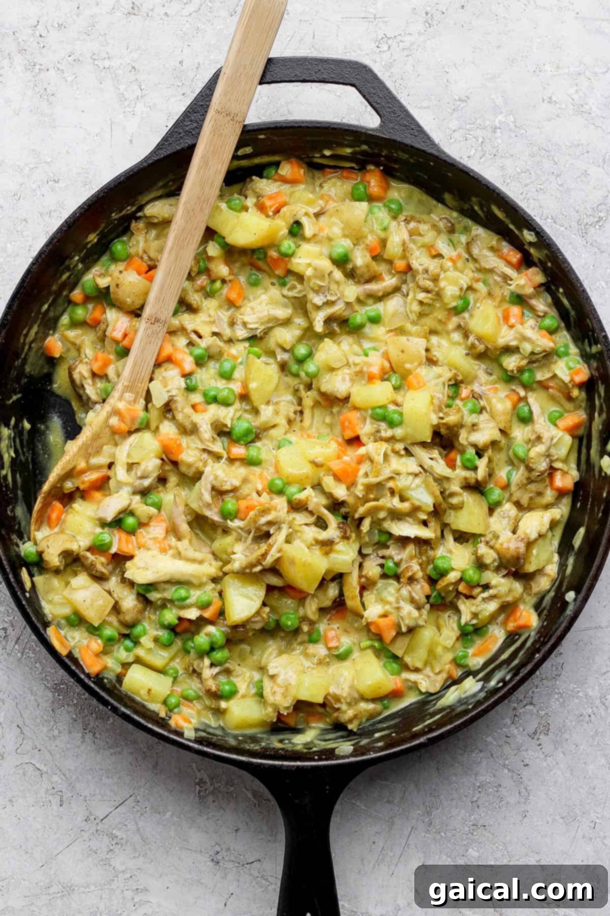 A skillet of cooked chicken, potatoes, carrots, and peas mixed in a creamy sauce, with a wooden spoon on the side.
