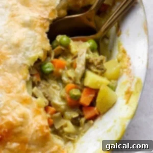 A close-up of a pot pie with a golden crust, showing a filling of chicken, peas, carrots, and potatoes, with two spoons partially inside.