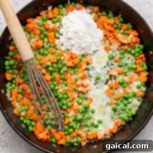 A skillet with sautéed carrots, peas, onions, butter, and a pile of flour, stirred with a whisk.