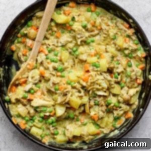 A skillet of cooked chicken, potatoes, carrots, and peas mixed in a creamy sauce, with a wooden spoon on the side.
