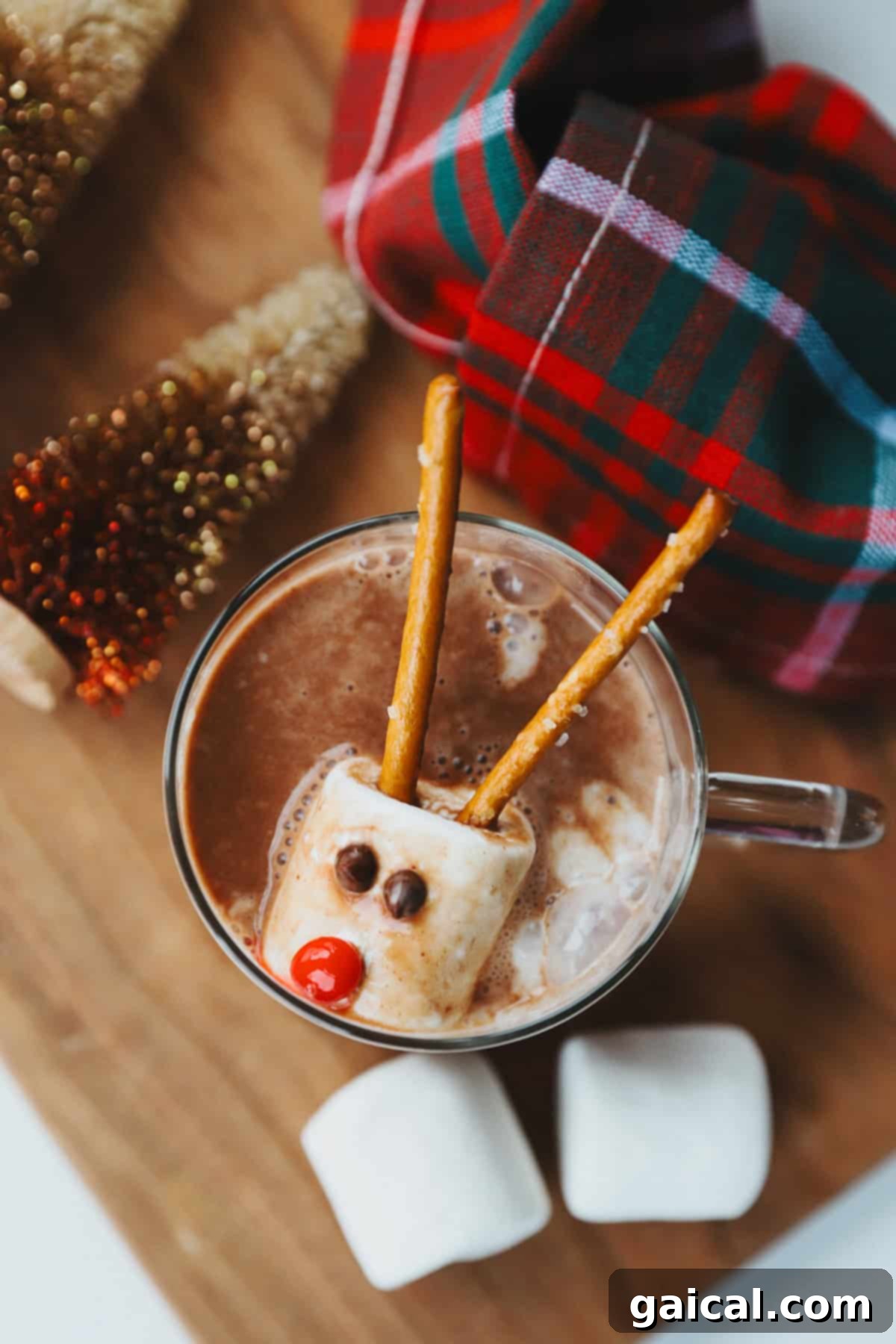 A festive cup of hot chocolate adorned with a marshmallow reindeer, complete with pretzel antlers, candy eyes, and a bright red candy nose, set against a cozy plaid cloth and surrounded by miniature Christmas trees.