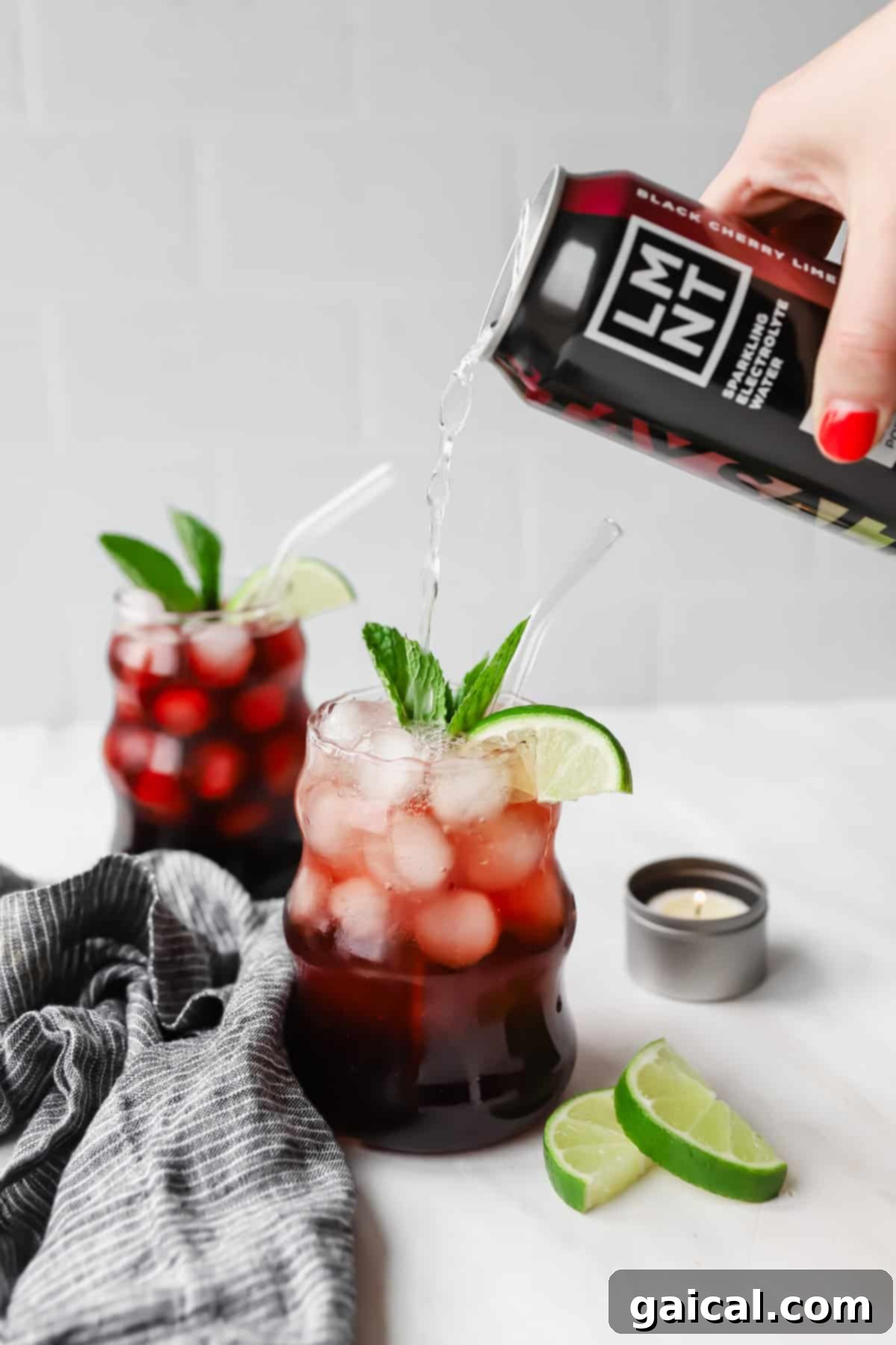 Nighttime Nectar 5 A hand pours a black cherry drink into a glass filled with ice, cherries, lime slices, and mint. Another similar drink is in the background, next to lime wedges and a small candle.