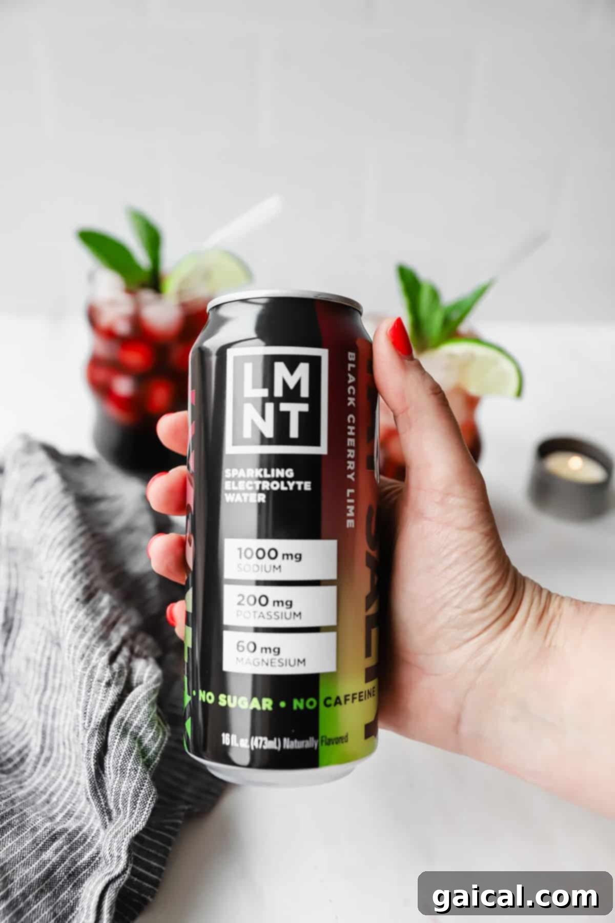 Nighttime Nectar 6 Hand holding a can of LMNT black cherry lime sparkling electrolyte water. Two glasses with red drinks and lime wedges are in the background.