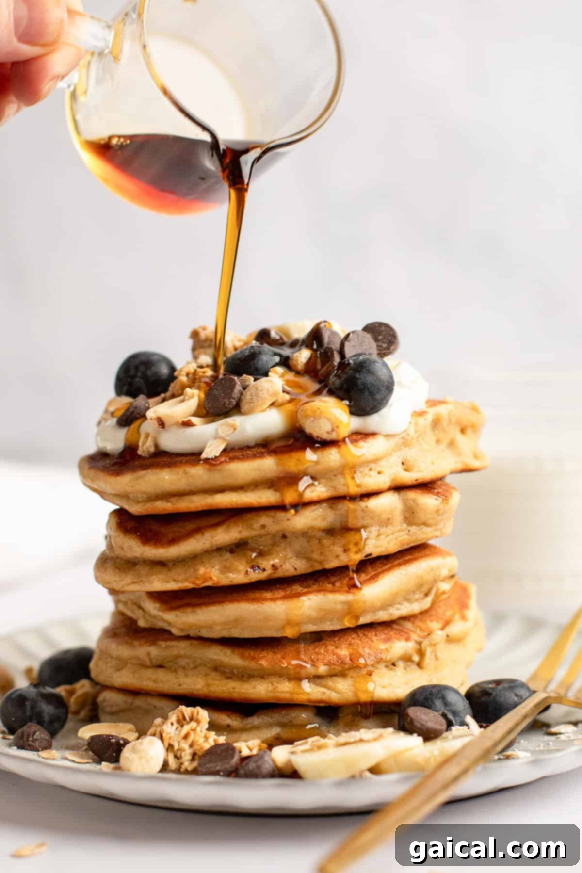 Power-Packed Pancakes 3 A beautifully arranged stack of golden-brown pancakes, generously topped with an array of fresh blueberries, crunchy chocolate chips, wholesome granola, and a sprinkle of nuts. A stream of rich maple syrup cascades over the stack, enhancing its appeal.