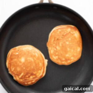 Power-Packed Pancakes 9 Two golden-brown pancakes cooking in a black non-stick frying pan on a white surface.