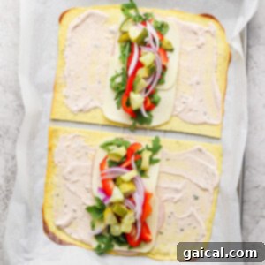 Two open-faced flatbread sandwiches on a baking sheet, with cheese, arugula, red onions, red peppers, and pickles, topped with a creamy spread, placed on parchment paper.