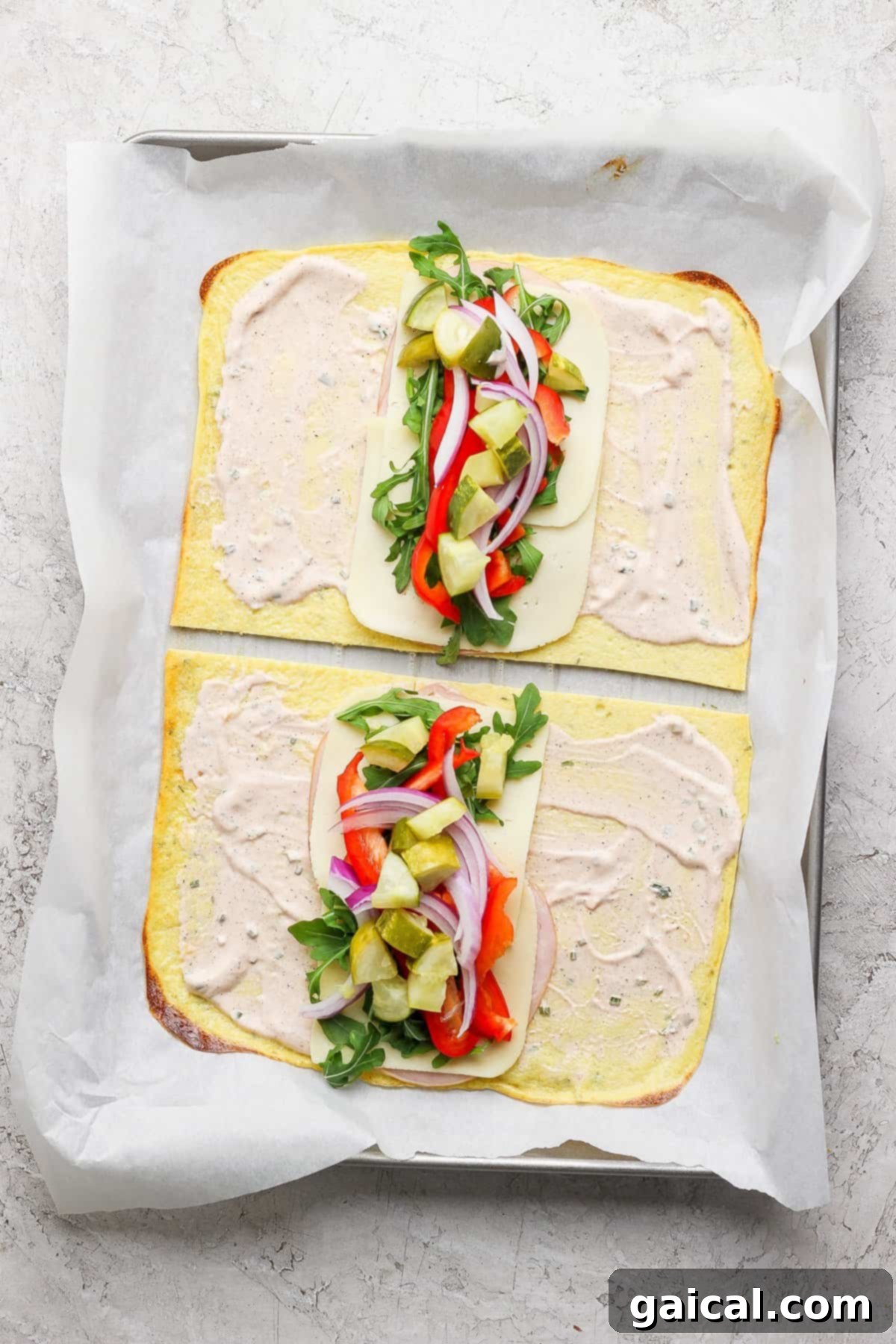 Protein-Packed Cottage Cheese Wraps 4 Two open-faced flatbread sandwiches on a baking sheet, with cheese, arugula, red onions, red peppers, and pickles, topped with a creamy spread, placed on parchment paper.