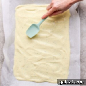 Hand spreading batter evenly on a baking sheet lined with parchment paper using a teal spatula.