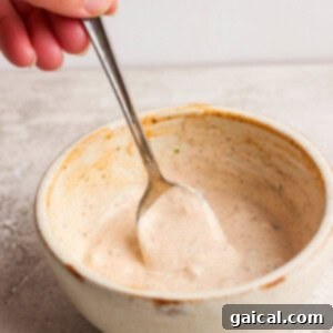 Hand holding a spoon in a ceramic bowl of creamy sauce on a gray surface.