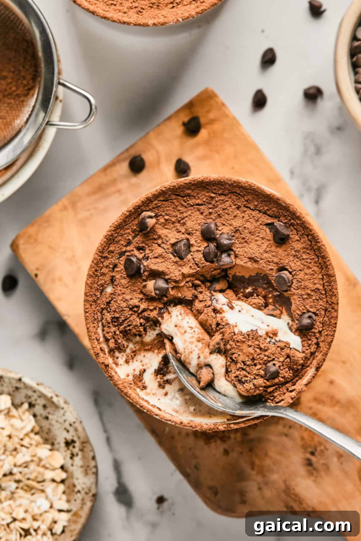 A bowl of Tiramisu Blended Oats topped with cocoa powder and chocolate chips, resting on a wooden board next to a sieve, loose oats, and scattered chocolate chips. A spoon is gently scooping a portion from the bowl.