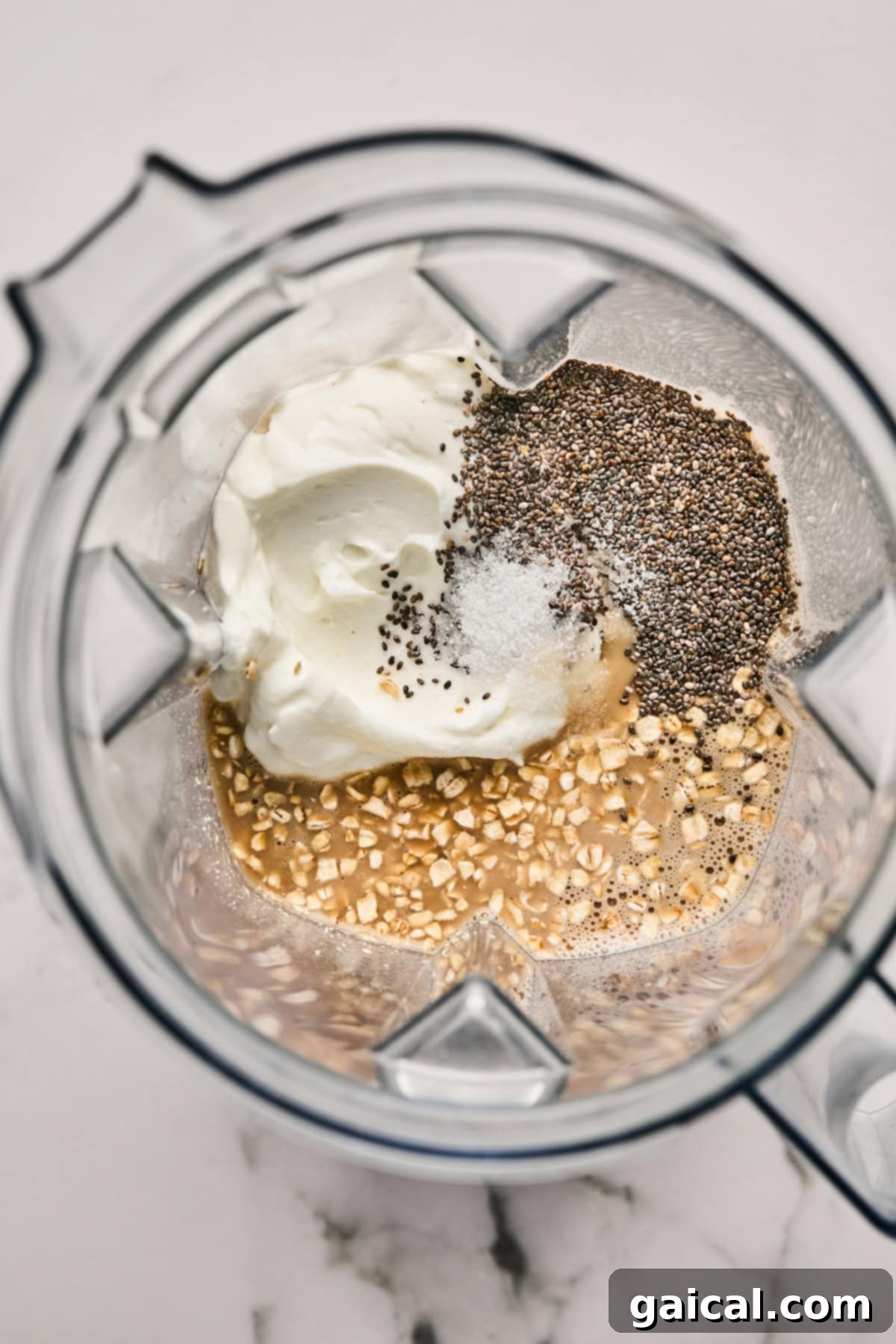 Ingredients for Tiramisu Blended Oats being added to a blender: creamy yogurt, small chia seeds, a pinch of salt, dry oats, and a liquid component.