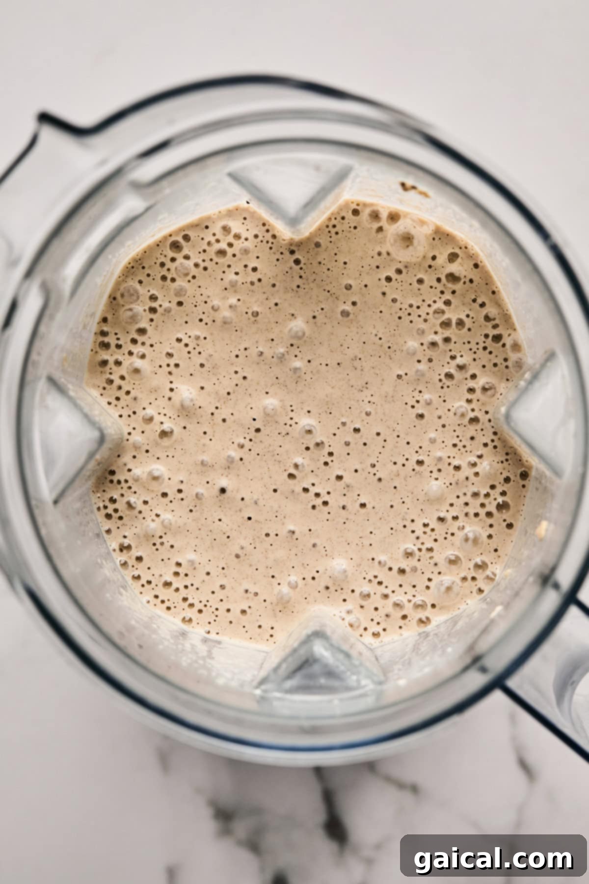 Top-down view of a blender filled with a smooth, frothy beige liquid, likely the blended oat mixture, sitting on a pristine white marble surface.