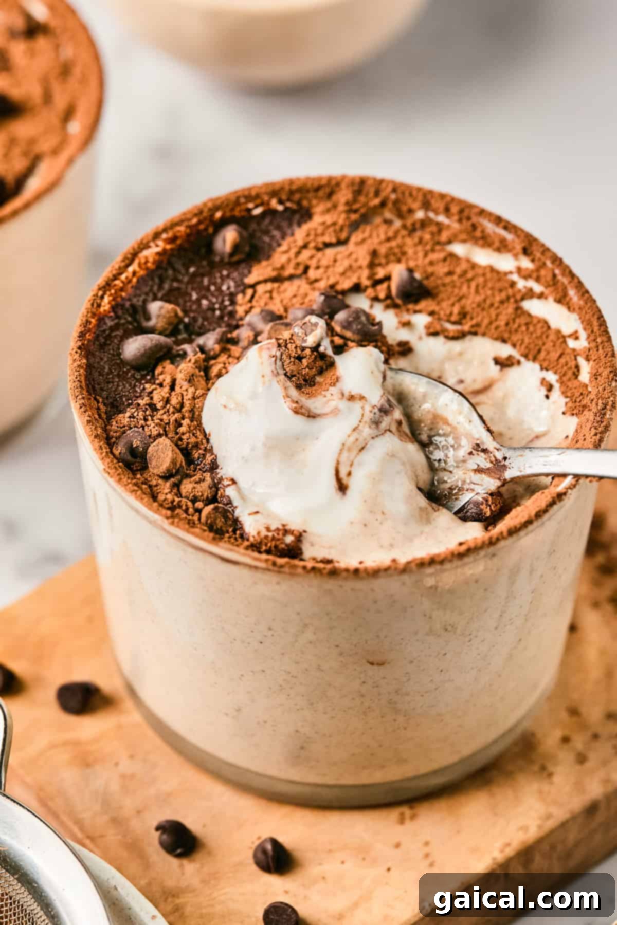 A beautifully presented serving of Tiramisu Blended Oats in a ceramic cup, generously topped with cocoa powder and mini chocolate chips, with a spoon having just scooped a portion.