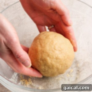 Protein-Packed Peanut Butter Twix Bars 7 Hands shaping a ball of dough in a clear glass bowl on a light-colored surface.