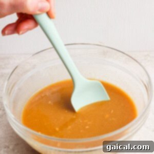 Protein-Packed Peanut Butter Twix Bars 9 A hand stirs brown gravy in a clear glass bowl with a light green spatula on a light surface.