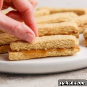 Protein-Packed Peanut Butter Twix Bars 10 A hand picks up a peanut butter sandwich cookie from a white plate. More cookies are visible in the background.