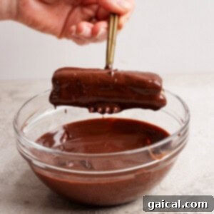 Protein-Packed Peanut Butter Twix Bars 11 A hand holds chopsticks dipping a chocolate-covered rectangular treat into a glass bowl of melted chocolate.