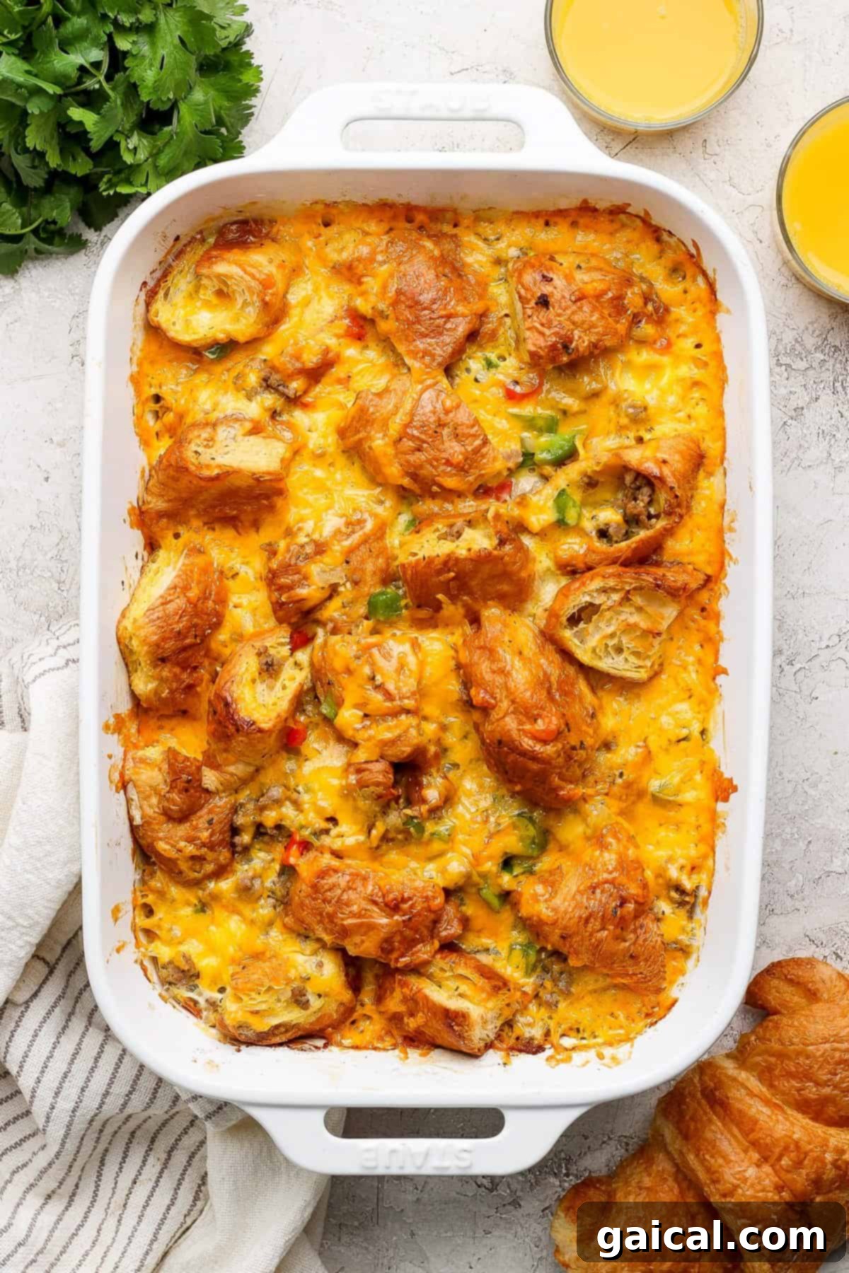 Buttery Croissant Egg Bake 2 A baked croissant egg bake in a white dish, topped with melted cheddar cheese and golden croissant pieces. Two glasses of fresh orange juice and a sprig of fresh herbs sit invitingly nearby.