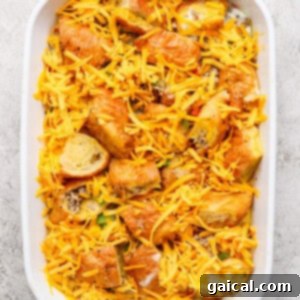 Buttery Croissant Egg Bake 11 A white baking dish filled with a partially baked casserole of bread cubes, sliced sausage, and vegetables, now topped with shredded cheese and ready for its final bake.