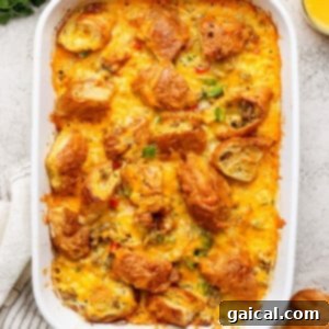 Buttery Croissant Egg Bake 12 A baked croissant egg bake in a white dish, topped with melted cheese and croissant pieces, resting before serving. Orange juice and fresh herbs are nearby.