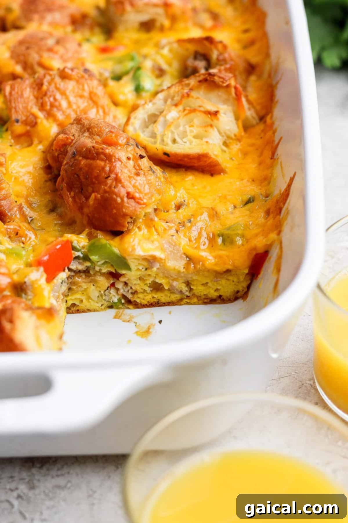 Buttery Croissant Egg Bake 4 A beautifully baked breakfast casserole in a white ceramic dish, showcasing rich melted cheese, colorful vegetables, and golden biscuit pieces, presented alongside two glasses of fresh orange juice.