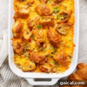 Buttery Croissant Egg Bake 5 A casserole dish filled with a baked croissant breakfast casserole, topped with melted cheese, savory sausage, and diced vegetables on a textured surface. A white striped cloth is partially visible.