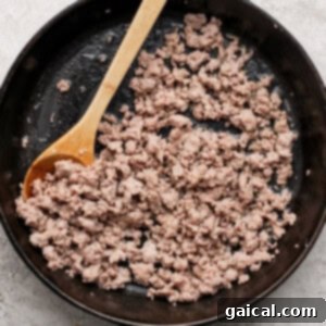 Buttery Croissant Egg Bake 7 Ground breakfast sausage cooking in a black cast-iron skillet, being broken apart with a wooden spoon.