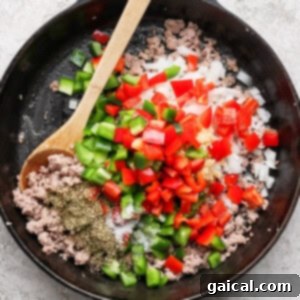 Buttery Croissant Egg Bake 8 A cast iron skillet showcasing a colorful mix of browned ground meat, chopped red and green bell peppers, diced onions, minced garlic, and herbs, stirred with a wooden spoon.