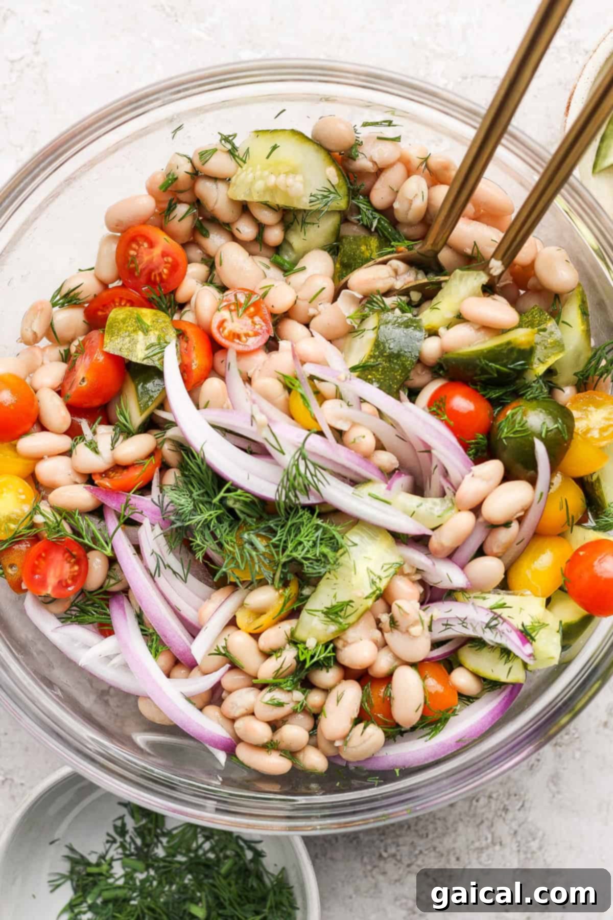 Zesty Dill Pickle White Bean Salad 2 A bowl of bean salad with white beans, cherry tomatoes, red onion, cucumber, fresh dill, and dressing, garnished with herbs, with serving utensils.
