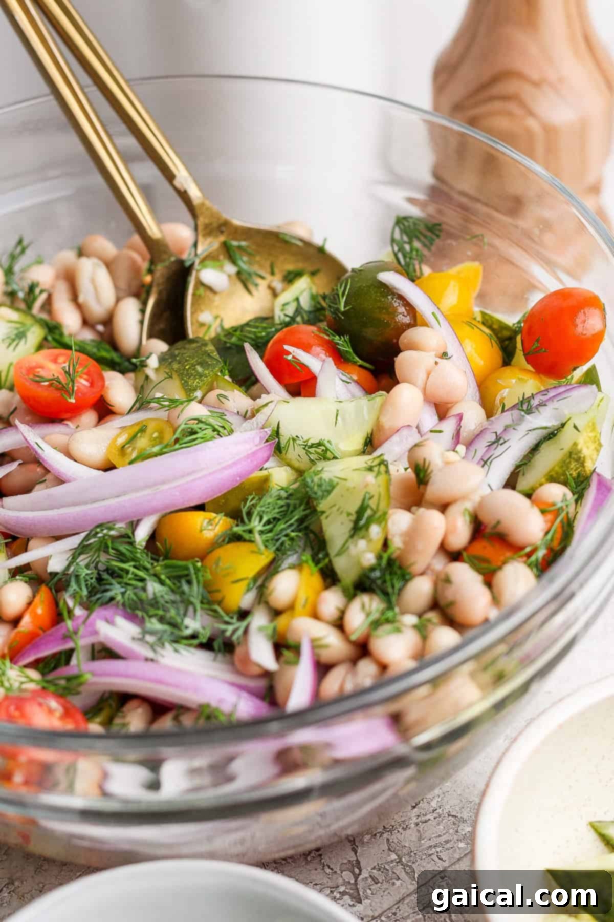 Zesty Dill Pickle White Bean Salad 5 A bowl of vegetable salad with cherry tomatoes, cucumber, white beans, red onion, and fresh dill, tossed with two gold utensils.