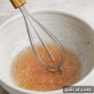 Zesty Dill Pickle White Bean Salad 8 A metal whisk rests in a white bowl filled with a light brown liquid, possibly a vinaigrette or sauce, set on a textured surface.