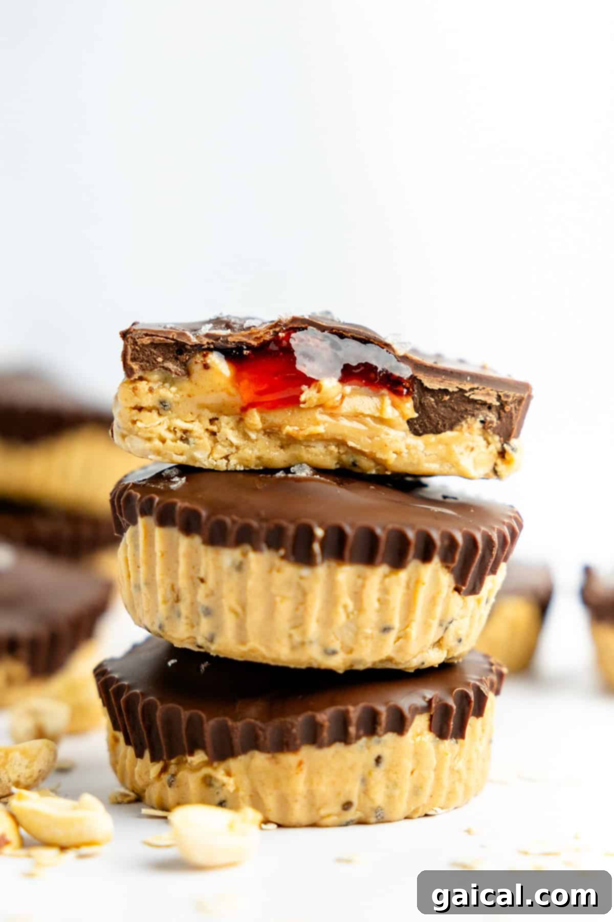 Three peanut butter and chocolate cups stacked, with a bite taken from the top one, revealing a vibrant red jam filling. Peanuts are scattered artfully around the cups, highlighting the key ingredient.