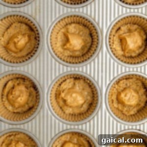 A muffin tray with paper liners, each filled with unbaked cookie dough and topped with a small dollop of peanut butter, showing the base layer being formed.