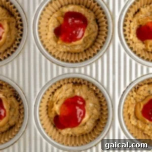 A muffin tin with unbaked muffin batter topped with vibrant red jam, nestled in paper liners, illustrating the filling stage.