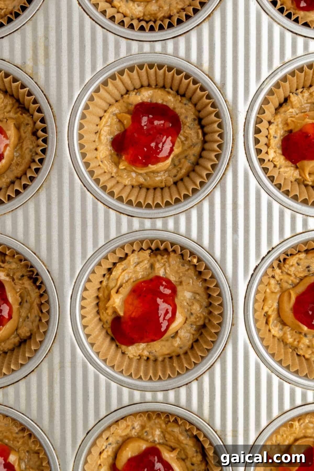 A muffin tin lined with paper wrappers, each filled with the unbaked oat cup batter and topped with a vibrant red dollop of jam, ready for the next step of preparation.