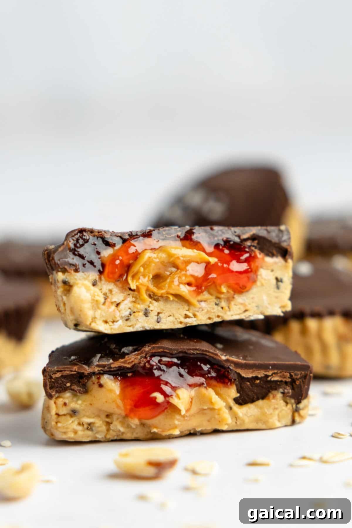 Two chocolate peanut butter cups, one cut in half to reveal layers of peanut butter and jelly filling. Surrounded by more peanut butter cups and scattered oats on a white surface.