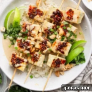 Tofu skewers with rice, garnished with lime wedges, cilantro, chopped nuts, and a drizzle of chili sauce, served in a white bowl.